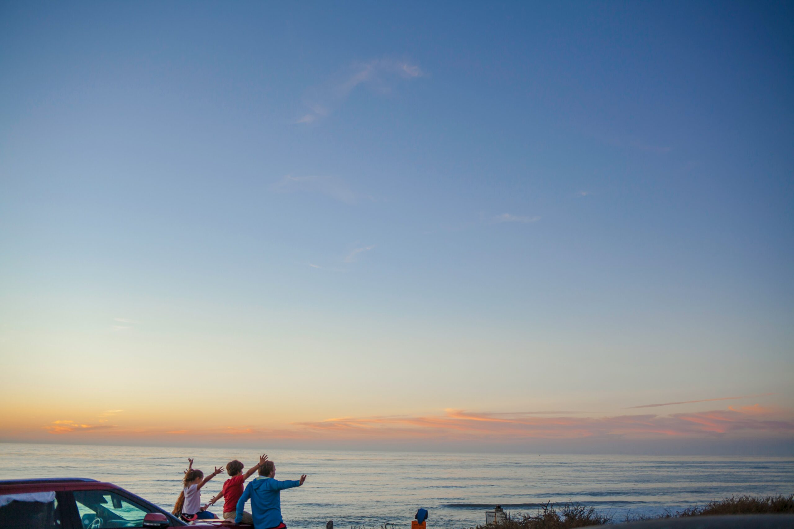 Family sitting on a rental car bonnet cheering at ocean sunset during a road trip